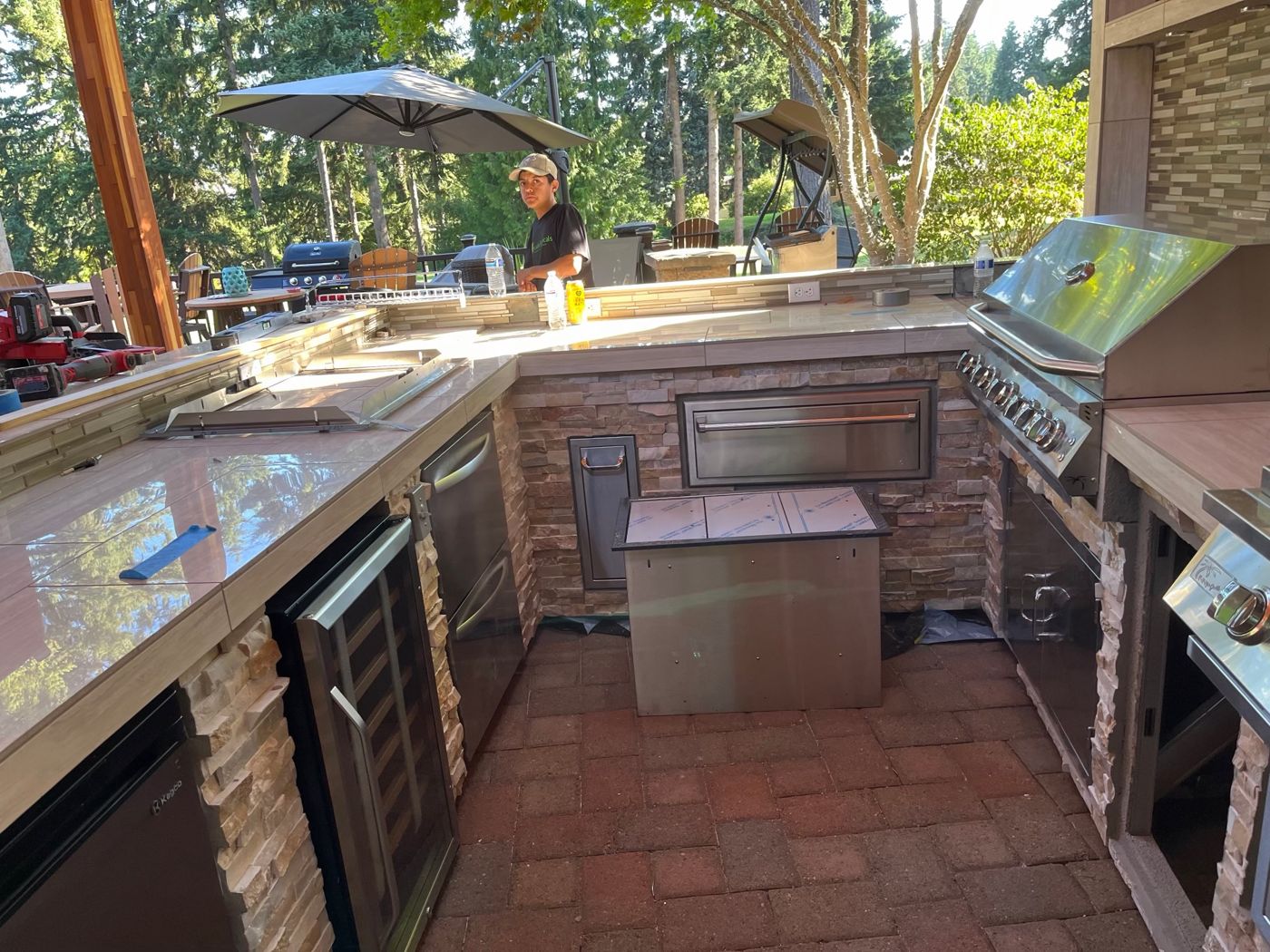 Outdoor kitchen being finished, with a stacked-stone base, tile countertops, built-in grill, fridges and storage